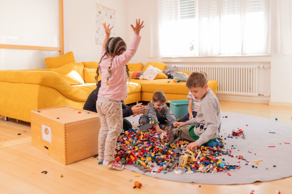 Vier Kinder sitzen und spielen mit vielen bunten Bausteinen auf einem runden Teppich vor einem gelben Sofa in einem hellen Wohnzimmer. Ein Kind steht und streckt vor Freude die Hände in die Höhe.