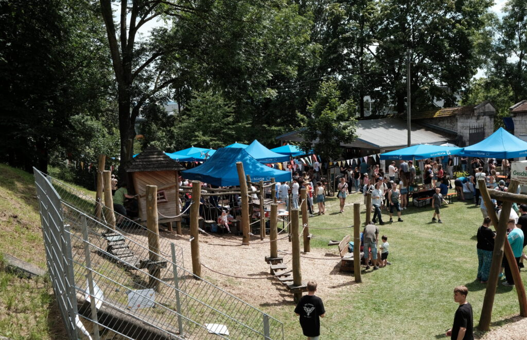 Spielplatz mit Klettergerüst und mehreren blauen Sonnenschirmen, umgeben von Menschen und Bäumen.