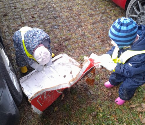 Zwei Kinder in bunten Overalls und Mützen ziehen an einem beschädigten Pappkarton. Eines trägt gelbe Gummistiefel, das andere pinke. Der Karton liegt auf dem Boden.