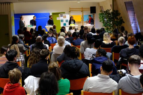 Eine Gruppe von Menschen sitzt in einem Raum und hört einer Präsentation zu. Im Vordergrund sind mehrere Personen mit Rücken zur Kamera zu sehen, während im Hintergrund eine Präsentationsfläche mit Plakaten und Tischen sichtbar ist.