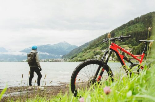 Ein Mountainbike in Rot und Schwarz steht im Vordergrund auf dem Gras. Im Hintergrund steht eine Person mit blauer Helm und Rucksack am Ufer eines Sees, umgeben von Bergen.