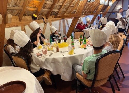 Gruppe von Kindern mit Kochmützen sitzt an einem runden Tisch, der mit einer weißen Tischdecke und Getränken gedeckt ist. Einige Kinder sind in Gespräche vertieft.